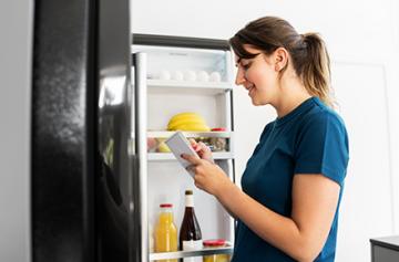Woman standing in front of an open refrigerator while making a list.