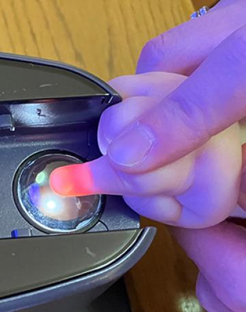 An adult's hand guides an infant's finger on a round clear scanner eye. The tip of the infant's finger appears orange due to the scanner light.