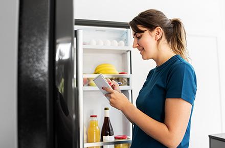 Woman standing in front of an open refrigerator while making a list.