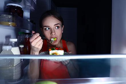 A woman standing in front of an open refrigerator in the dark eating.