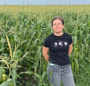 Una mujer con una camiseta negra y pantalones vaqueros está parada frente a un campo de tallos de maíz.