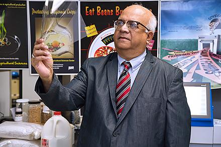 A man in a suit jacket, tie, and glasses holds up and looks at a clear rectangular sheet - Atanu Biswas inspects a piece of bioplastic in his lab.