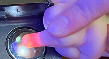 An adult's hand guides an infant's finger on a round clear scanner eye. The tip of the infant's finger appears orange due to the scanner light.