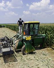 Three people preparing tractor-mounted sprayer equipment in a cornfield.