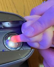 An adult's hand guides an infant's finger on a round clear scanner eye. The tip of the infant's finger appears orange due to the scanner light.