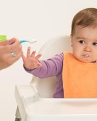 A baby in a high chair looks unhappily at some food that is being offered by a nearby adult holding a bowl and spoon.